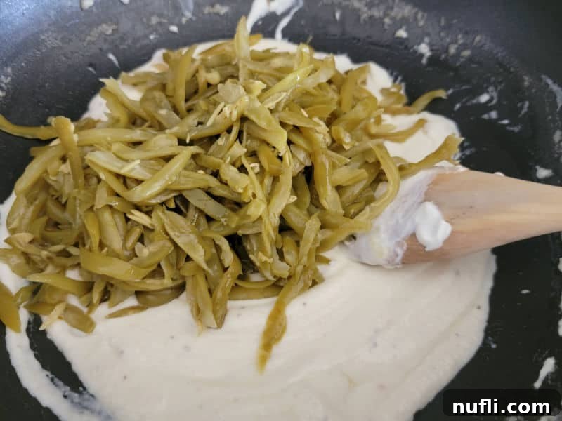 Drained green beans being added to and stirred into the rich sour cream sauce in a black skillet.