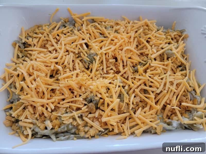 Shredded cheddar cheese covering the cornflake-topped green beans in a white casserole dish.