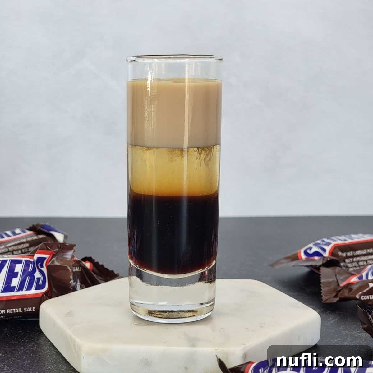 Frangelico Creations 6 Layered Snickers Shot on a white coaster surrounded by Snickers Chocolate Bars