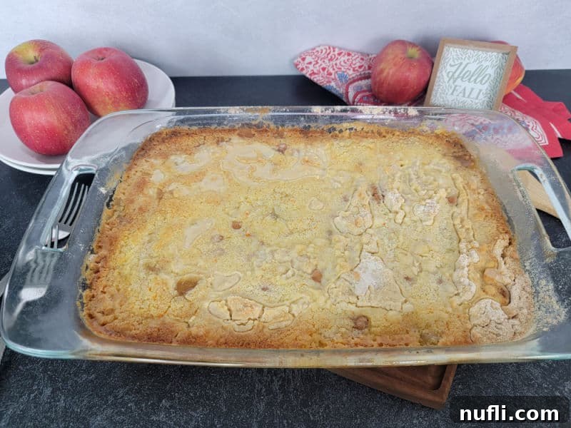 A freshly baked Caramel Apple Dump Cake, golden brown and bubbling, cooling in its glass baking dish on a wooden surface with a decorative cloth towel.
