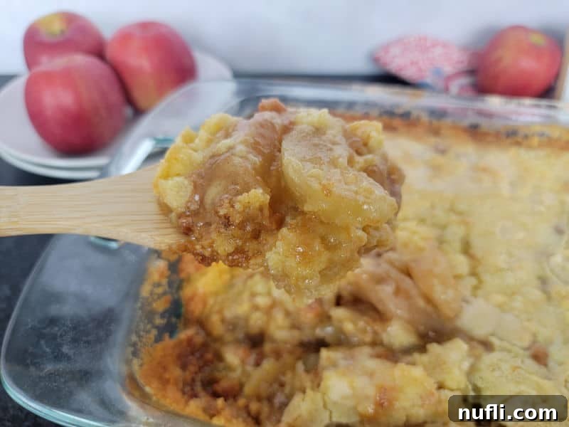 A wooden spoon scooping a portion of Caramel Apple Dump Cake from a glass casserole dish, showing the delicious layers of baked apples, melted caramel, and golden cake, with fresh apples in the background.