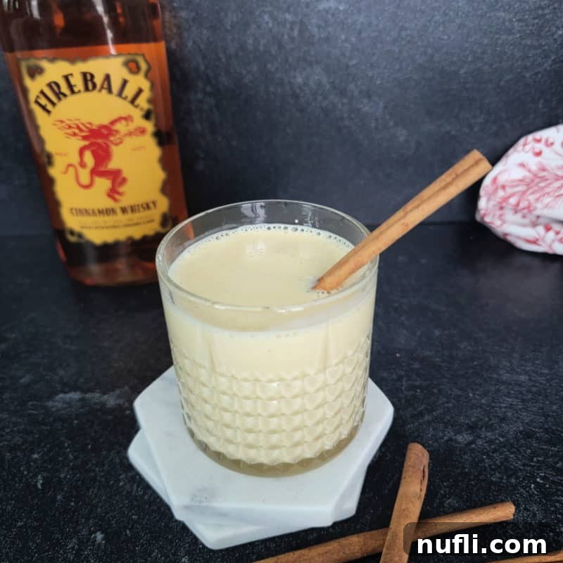 Fireball Eggnog in a crystal glass with cinnamon stick garnish in front of a bottle of Fireball Cinnamon Whiskey