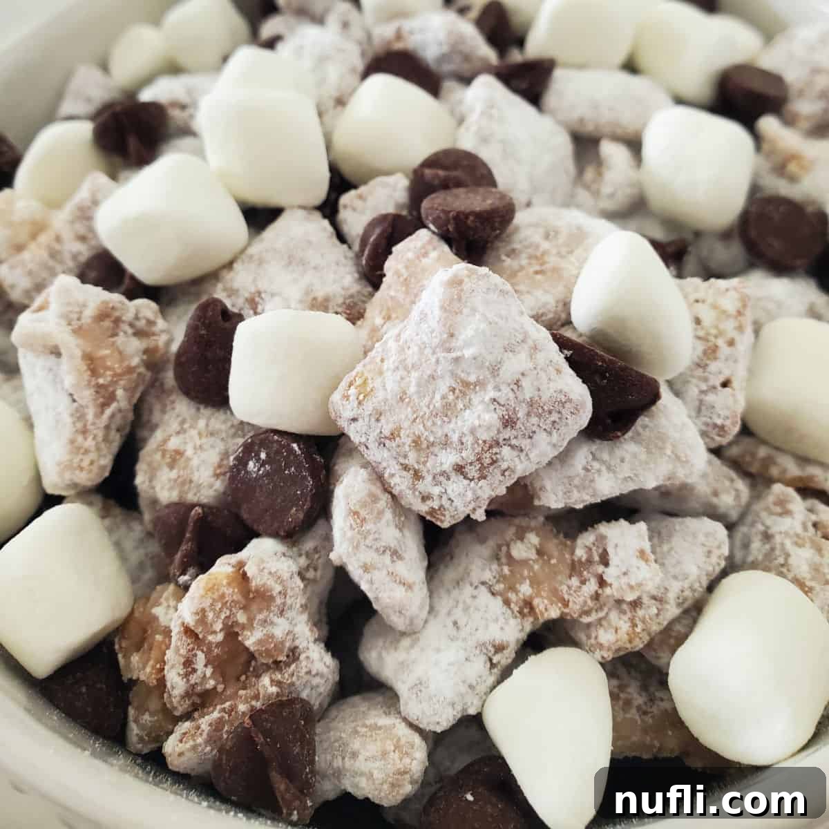 Hot chocolate muddy buddies piled in a white bowl with mini marshmallows and chocolate chips