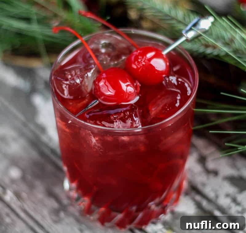 A festive red cherry cocktail, garnished with two maraschino cherries, artfully placed beside lush greenery.