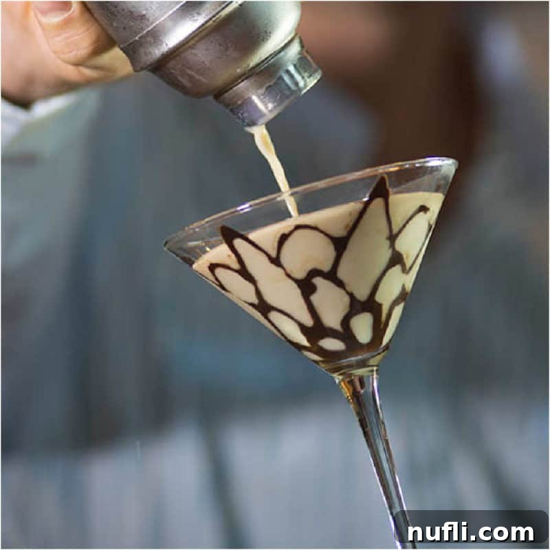 A Tiramisu Martini being skillfully poured from a cocktail shaker into a martini glass, which features an artistic chocolate design.