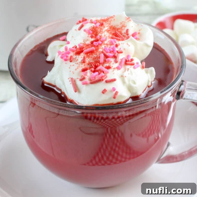 A rich Red Velvet Hot Chocolate in a clear glass mug, lavishly topped with whipped cream and a sprinkle of pink decorations.
