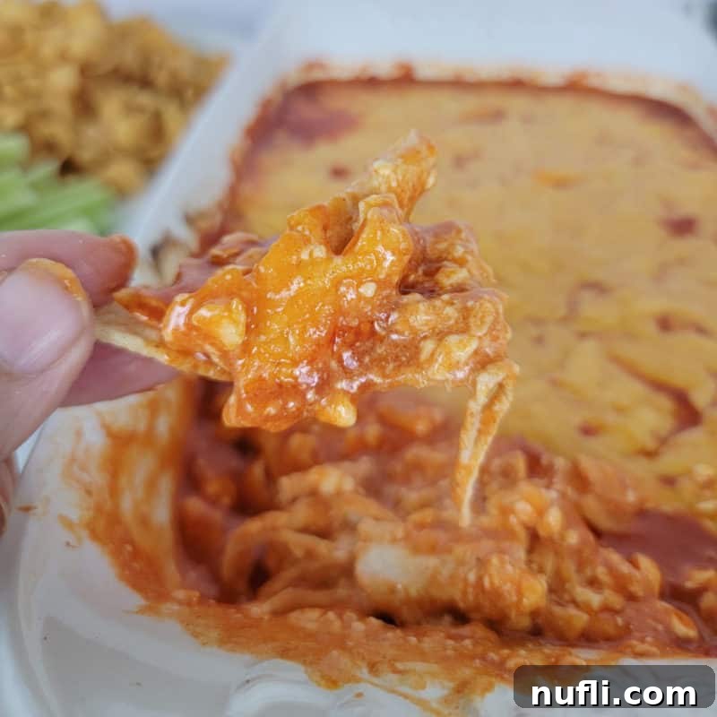 A chip laden with Easy Frank’s Buffalo Chicken Dip, held above a full baking dish of the piping hot dip