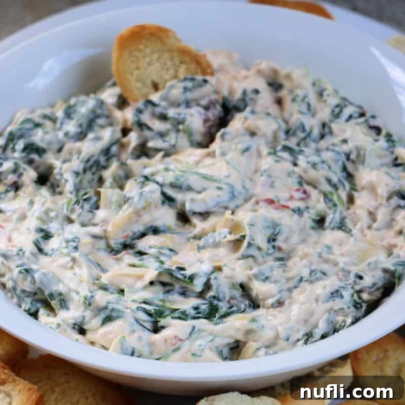Luxurious Creamy Spinach Artichoke Dip in a white bowl, ready to be enjoyed with a freshly sliced baguette
