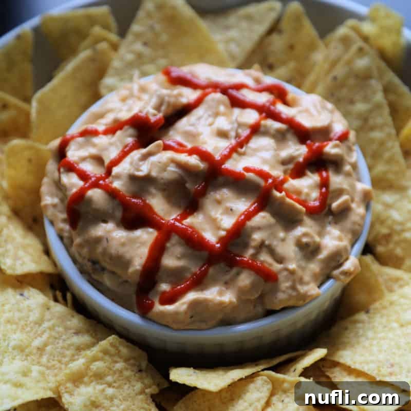 Cheesy Crock Pot Sriracha Chicken Dip, garnished with a drizzle of sriracha, served in a white bowl and surrounded by tortilla chips