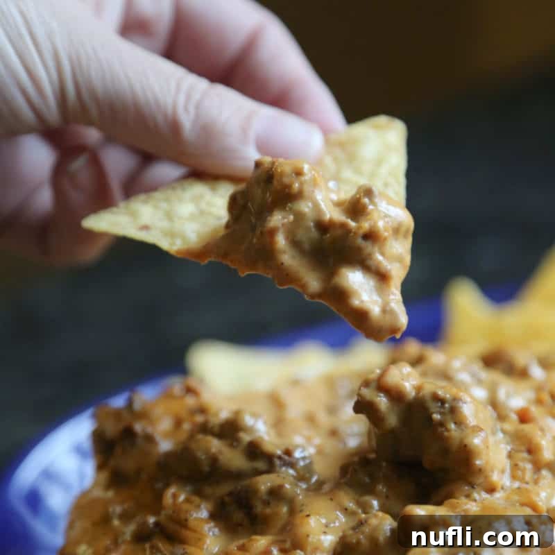 A tortilla chip generously loaded with Meat Lovers Queso, hovering above a bowl brimming with the rich, cheesy dip