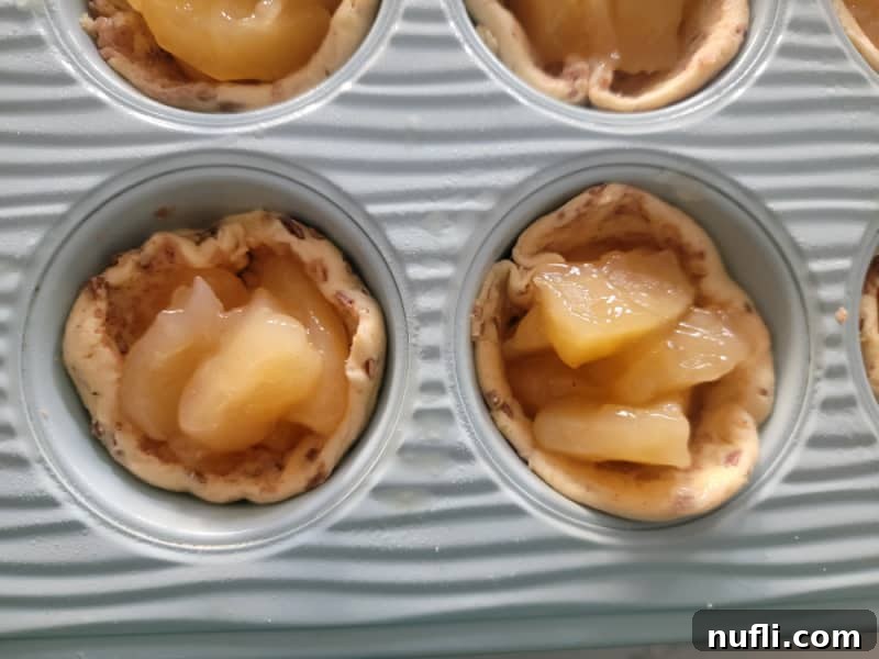 2-Ingredient Cinnamon Roll Apple Pie Bites 6 Unbaked Cinnamon Roll Apple Pie cups in a blue muffin tin, filled with apple pie filling.