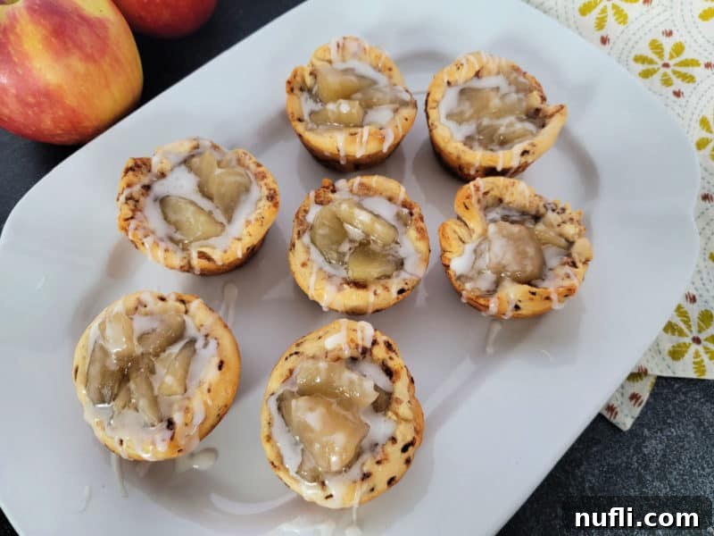 2-Ingredient Cinnamon Roll Apple Pie Bites 8 Beautifully arranged Cinnamon Roll Apple Pie Cups on a white platter, with fresh apples blurred in the background.