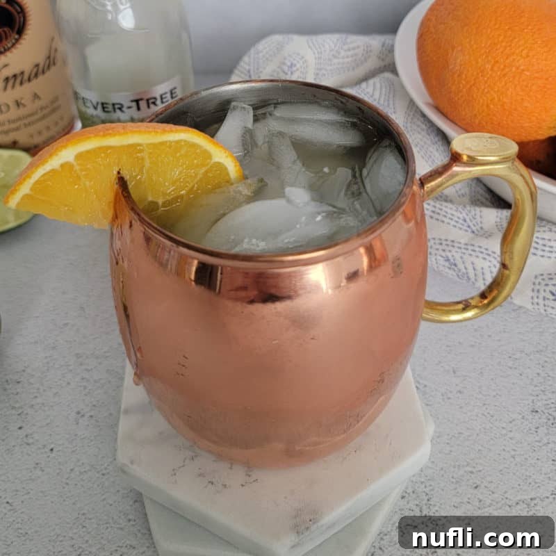 A vibrant Orange Moscow Mule served in a classic copper mug, artfully arranged next to a rustic bowl filled with fresh oranges, a bottle of zesty ginger beer, and a bottle of smooth vodka, showcasing its key ingredients.