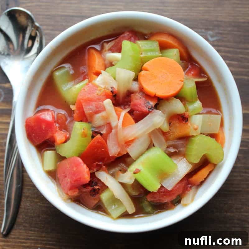 A hearty bowl of WW Vegetable Soup, brimming with colorful carrots, celery, onions, and diced tomatoes, with a spoon nestled beside it.