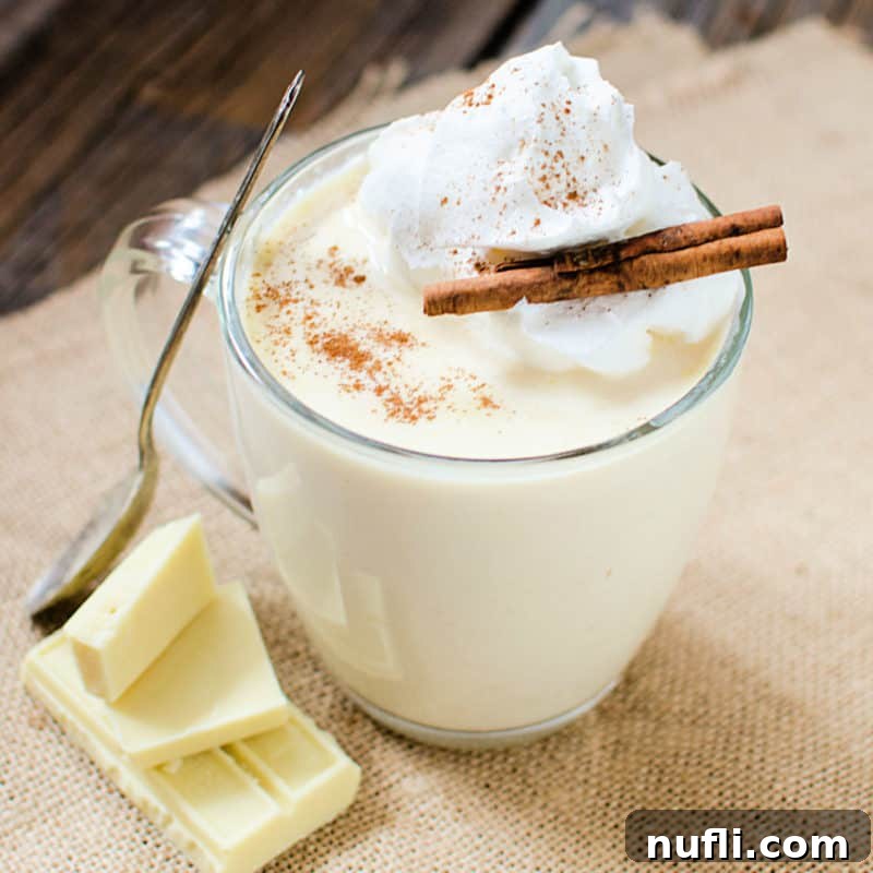 white hot chocolate in a glass coffee mug with a silver spoon, white chocolate pieces, and cinnamon sticks