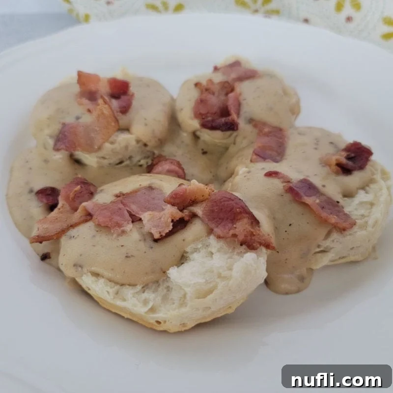 Rich Bacon Gravy generously poured over warm biscuit halves on a white plate, with a cloth napkin in the background.