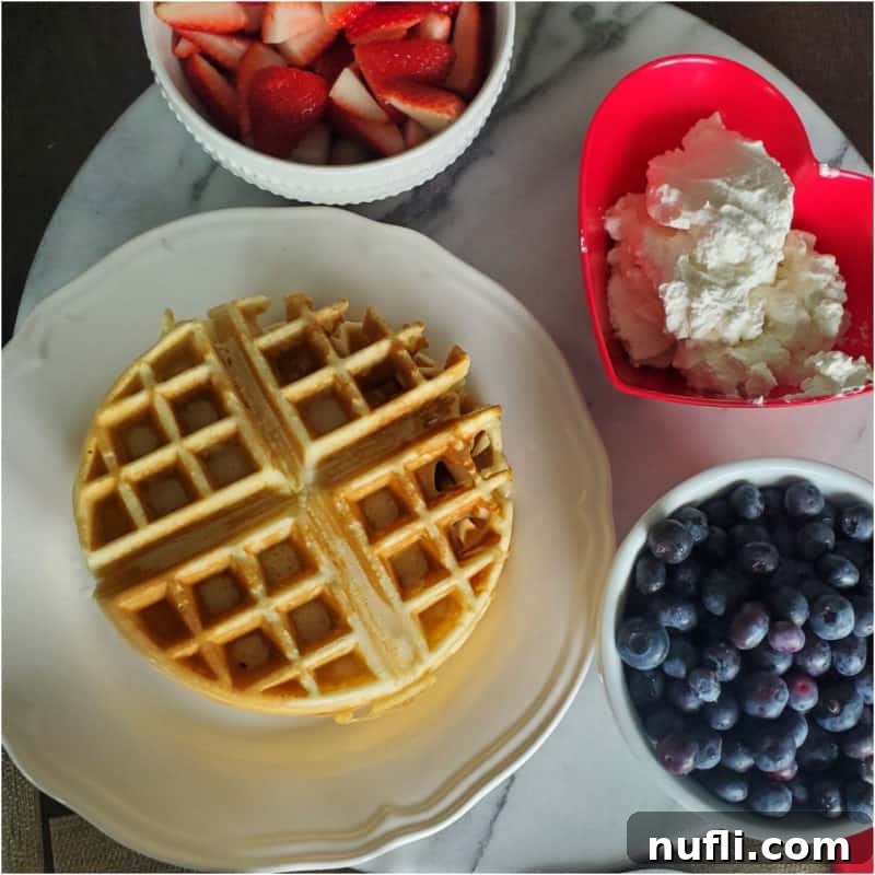 A perfectly golden Bisquick Waffle on a white plate, accompanied by fresh strawberries, whipped cream, and a bowl of blueberries.