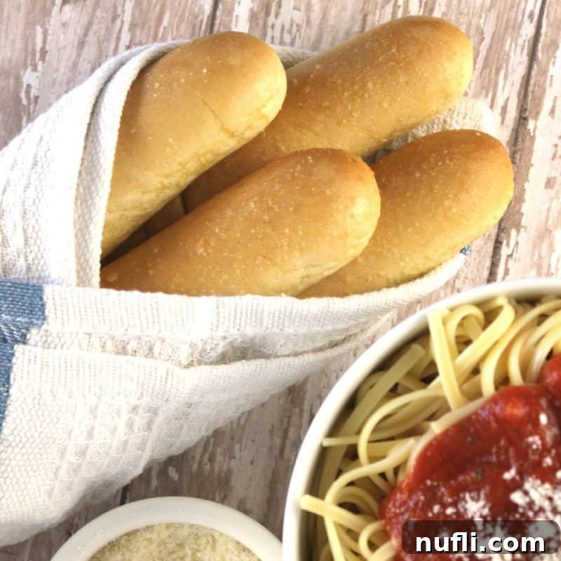 Freshly baked Olive Garden breadsticks, perfectly garlicky and golden, wrapped in a cloth next to a bowl of spaghetti, ready for dipping.