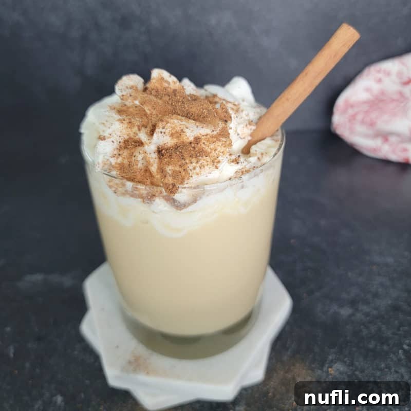 Eggnog Mudslide cocktail served in a glass on a white coaster, garnished with fluffy whipped cream and a sprinkle of nutmeg, alongside a cinnamon stick.