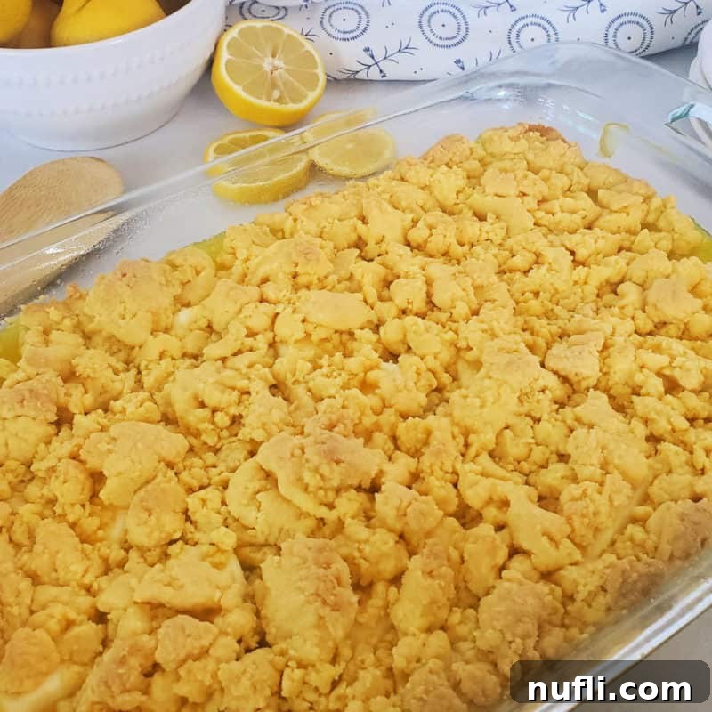 A glass casserole dish filled with bubbling Lemon Dump Cake, with a bowl of fresh lemons artfully blurred in the background.