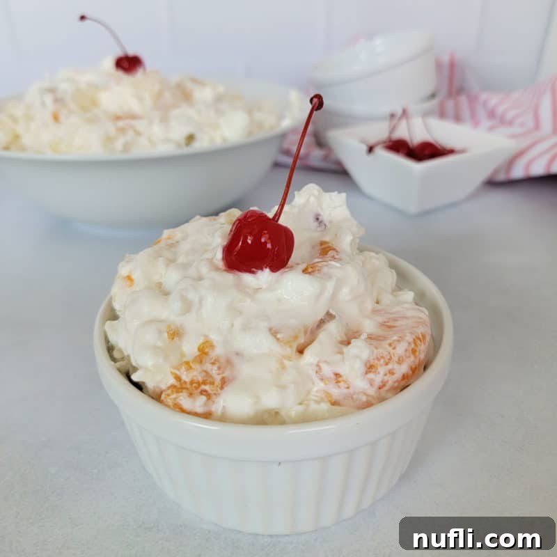 A small ramekin holds a portion of classic Ambrosia Salad, adorned with a single maraschino cherry for garnish.