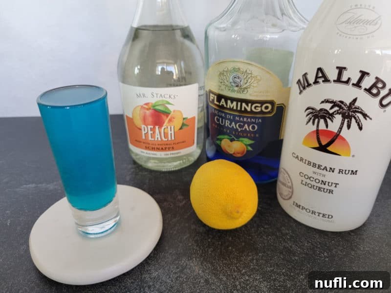 The key ingredients for a Blue Balls Shot: a shot glass of blue liquid, surrounded by bottles of peach schnapps, Blue Curacao, Malibu Coconut Rum, and a fresh lemon slice, all neatly arranged on a white surface.