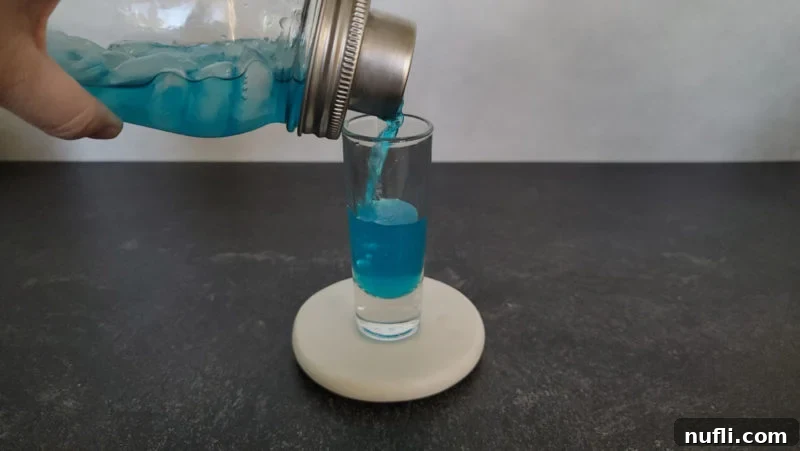 A beautiful blue cocktail shot being poured from a mason jar-style cocktail shaker into a classic shot glass, highlighting the rich blue color and inviting texture.