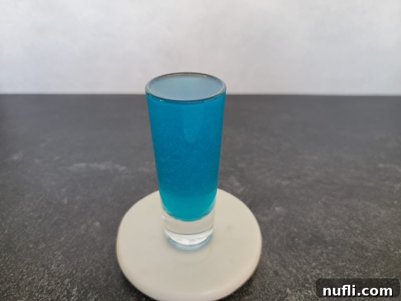 A vibrant blue cocktail shot, garnished with a fresh lemon slice, sitting elegantly on a white coaster, exuding a refreshing and inviting appeal ready to be enjoyed.