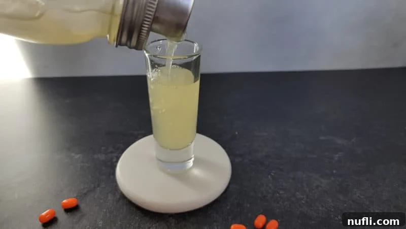A skilled hand pouring the bright yellow liquid of an Orange Tic Tac Shot from a cocktail shaker into a small shot glass, with orange Tic Tacs scattered nearby, highlighting the final step of preparation.