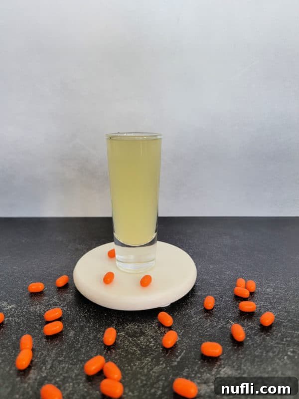 A vibrant yellow shot, the Orange Tic Tac Shot, presented elegantly on a white coaster and surrounded by numerous orange Tic Tac candies, highlighting its inviting color and theme.
