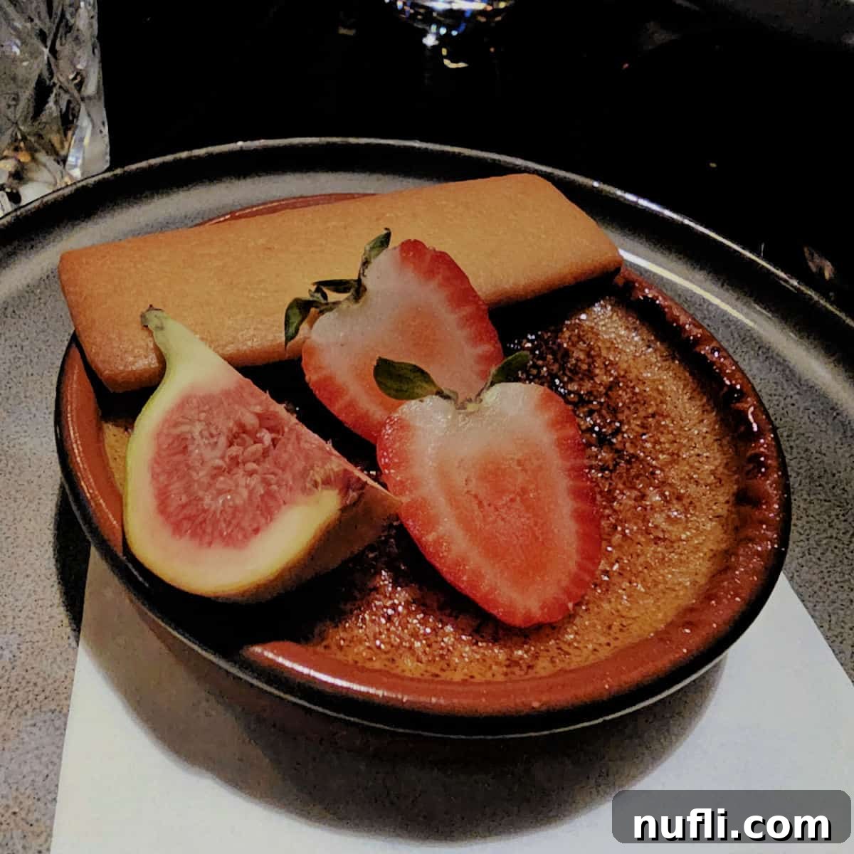 A decadent maple crème brûlée, garnished with fresh strawberries and a fig, served alongside a delicate maple cookie