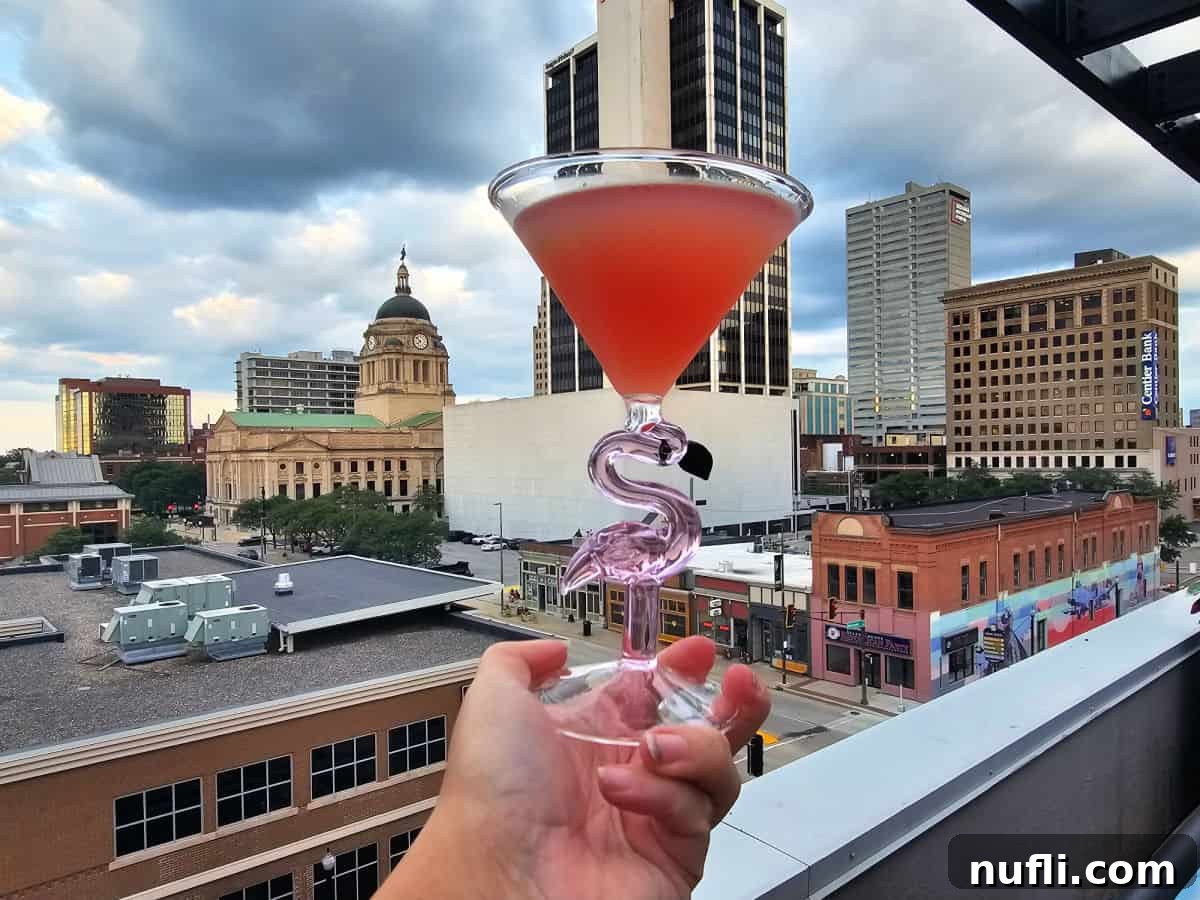 Hand holding a pink drink in a flamingo glass with the Fort Wayne skyline in the background