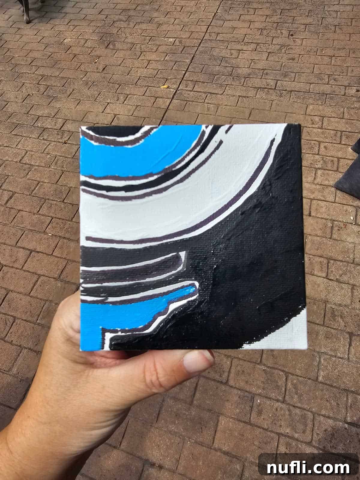 hand holding a small square canvas with white, blue and black painting