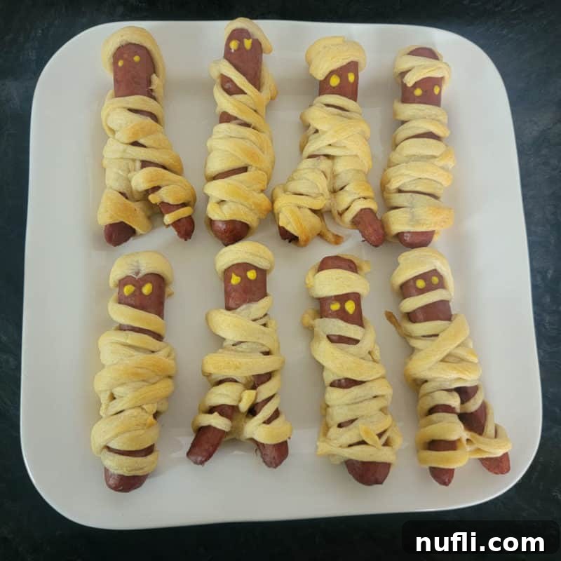 Delightful Mummy Hot Dogs arranged on a pristine white plate, showcasing their golden-brown crescent roll bandages and tiny mustard eyes, ready for a Halloween celebration.