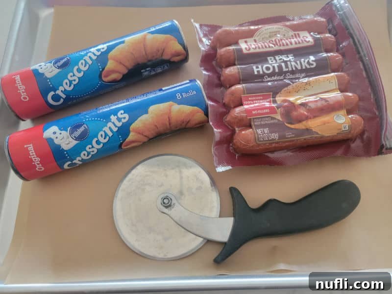 Raw ingredients for mummy hot dogs: crescent rolls, Johnsonville beef hot links, and a pizza cutter on a parchment-lined baking sheet, awaiting transformation.