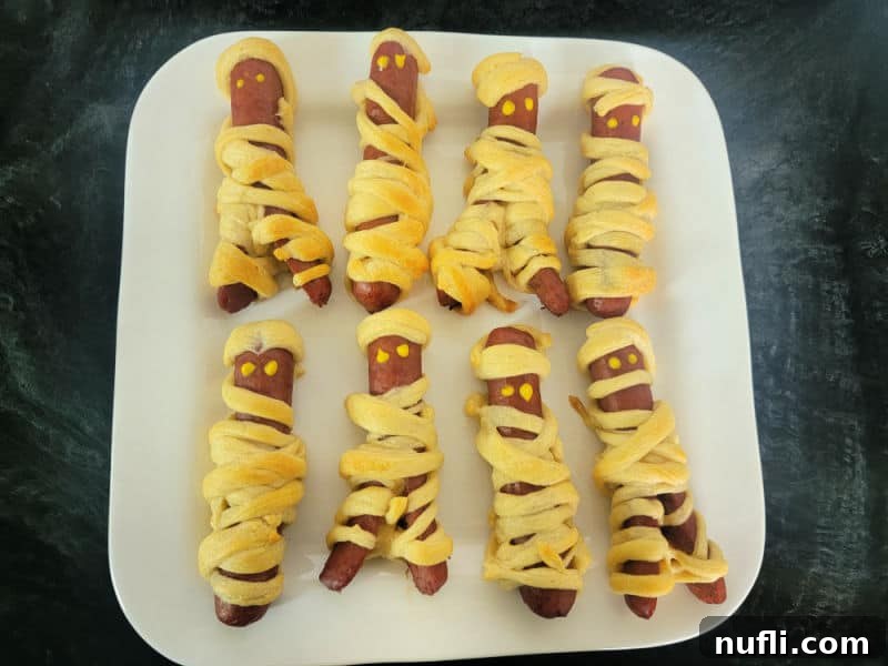 Baked crescent-wrapped mummy hot dogs resting on a white plate, showing off their golden-brown bandages before the final touch of eyes.