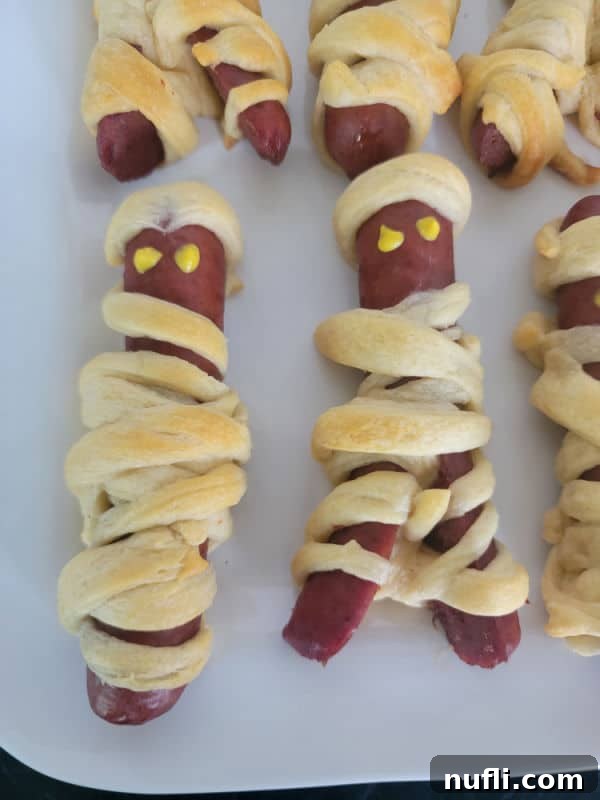 A close-up shot of several mummy hot dogs on a white plate, now complete with adorable mustard eyes, ready to be devoured at a Halloween gathering.