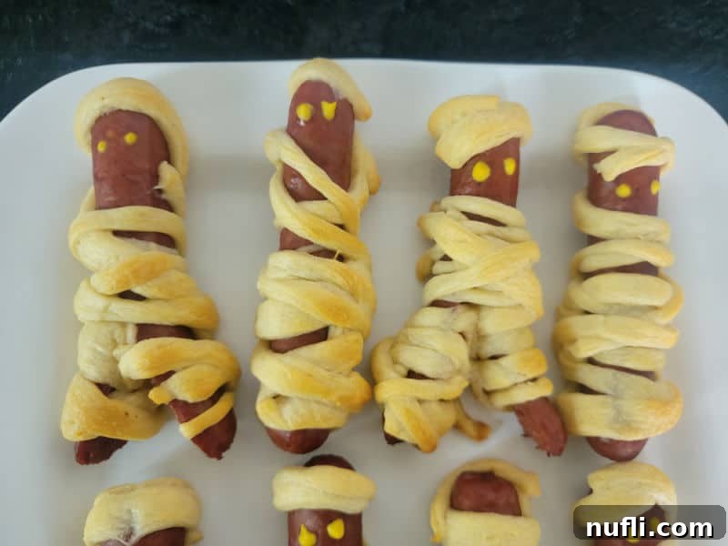 A festive plate of Mummy Hot Dogs, with some featuring distinct mustard eyes, arranged invitingly on a white surface, ready to be enjoyed as a Halloween snack or meal.