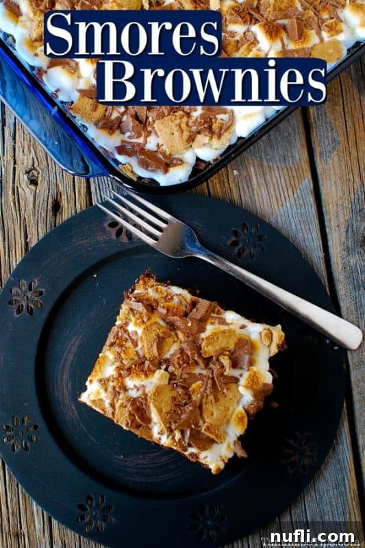 Ultimate S'mores Brownies 2 A delicious S'mores brownie square on a blue plate with a fork next to it. In the background, a baking pan filled with more brownies rests on a rustic wood surface.