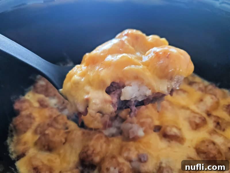 Serving spoon holding a portion of cheesy tater tots and ground beef over a slow cooker bowl, highlighting the layers.