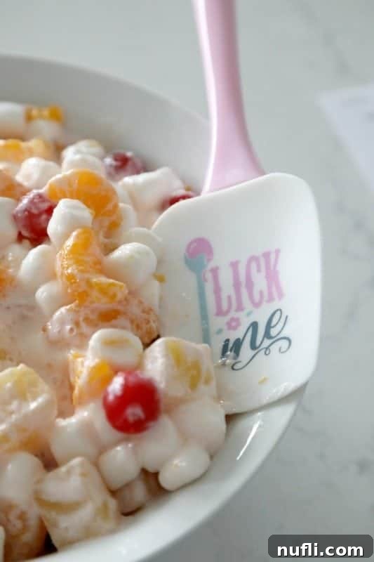Close-up of a creamy and vibrant fruit salad, showcasing pineapple chunks, mandarin oranges, and maraschino cherries mixed with mini marshmallows.
