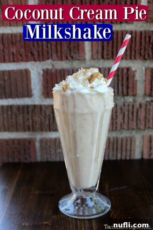 A tall, clear glass filled with a creamy white coconut cream pie shake, garnished with whipped cream and graham cracker crumbs, stands on a rustic brick background. A pink and white striped straw is inserted into the shake.