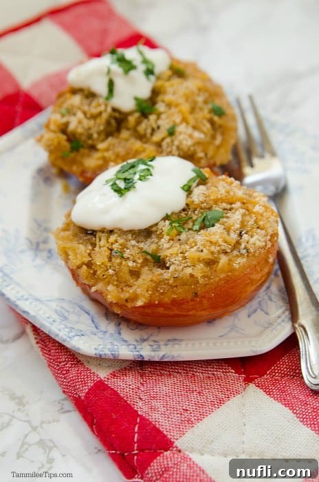 A white plate with a silver fork, holding a cheese-stuffed tomato adorned with sour cream and basil/parsley, resting on a red and white checkered oven mitt.