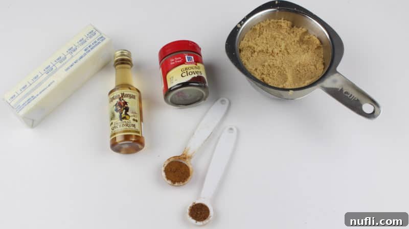 A stick of butter, spiced rum bottle, ground cloves, brown sugar, and more spices on a white counter