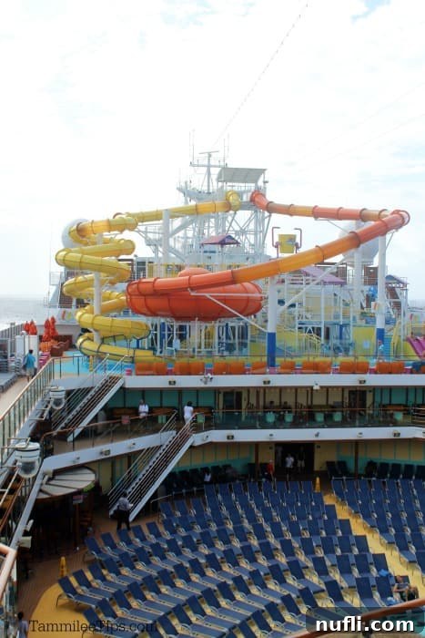 Carnival Waterworks: Your Ultimate Summer Splash 3 Panoramic view of the lively pool deck on the Carnival Breeze, showcasing two prominent waterslides and abundant lounge chairs, under a clear sky.