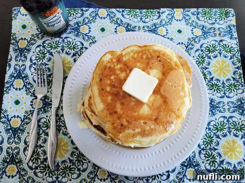 Homemade Cracker Barrel Buttermilk Pancakes 4 cracker barrel pancakes on a white plate with a square of butter, next to a fork, knife, and bottle of syrup