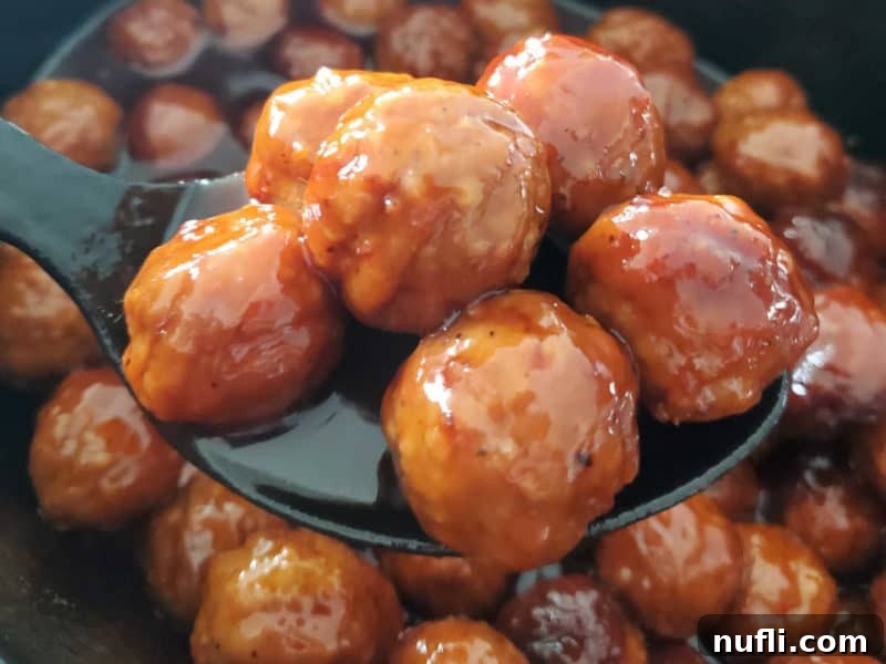 Easy & Delicious 3-Ingredient Grape Jelly Meatballs 6 A large serving spoon holds a portion of grape jelly meatballs, dripping with sauce, poised over a black slow cooker.
