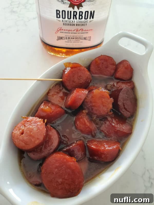 Close-up of sliced kielbasa coated in a rich, glossy bourbon glaze, served in a white bowl with a bottle of bourbon in the background.