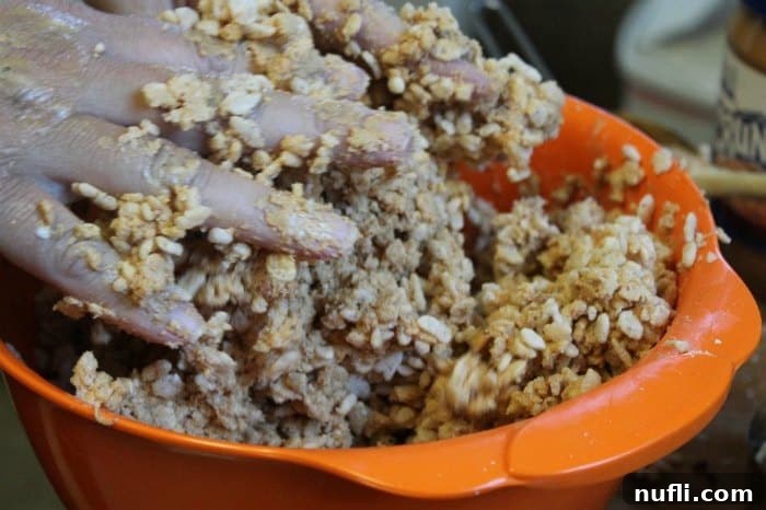 Hands actively mixing the peanut butter and Rice Krispies mixture in an orange bowl, demonstrating the hands-on approach for best results.