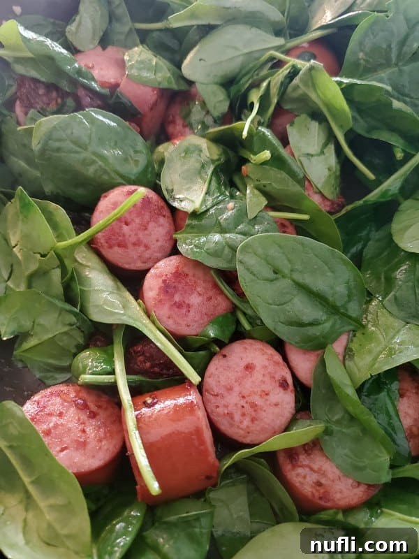 Close-up of vibrant green baby spinach leaves and slices of golden-brown kielbasa sausage in a pan, showcasing their readiness to be combined with other ingredients.